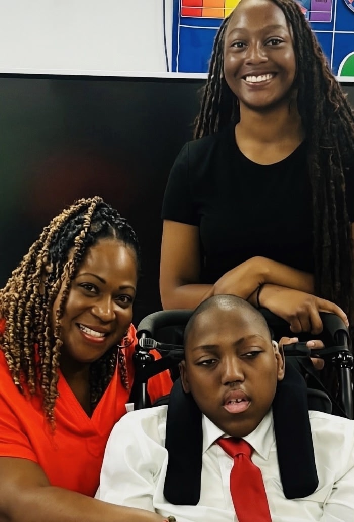 Beaming Crystal hugging Dwight with Kiyah leaning on the back of his wheelchair, smiling. Dwight is wearing a red tie that matches Crystal's dress.